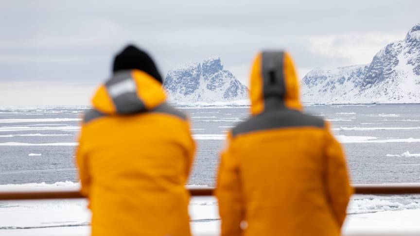 Two people stand on a ship with their backs to the camera, wearing their Quark Expeditions parkas, looking out onto an icy landscape