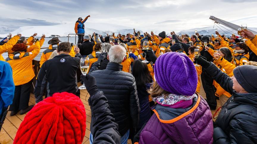 Guests on the deck of a polar expedition ship, wearing Quark Expedition parkas, gathered around an expedition leader while everyone raises a glass of champagne in celebration against a backdrop of ice