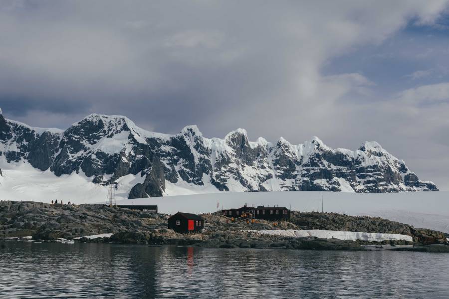 Water-level view of Port Lockroy captured from a zodiac boat. Building of the station stand out due to their red roof and guests in yellow parkas can be seen entering the post office and gift shop building.