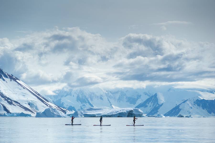 Guests go standup paddleboarding n the Antarctica Peninsula. Snow covered mountains are seen in the distance.