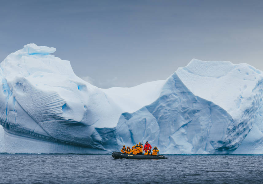 Zodiac boat in front of iceberg. Guests in yellow parkas can be seen taking photos while their Expedition Guide, in red, drives the boat.