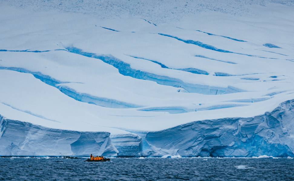 Zodiac boat in front of iceberg. Guests in yellow parkas can be seen taking photos while their Expedition Guide, in red, drives the boat.