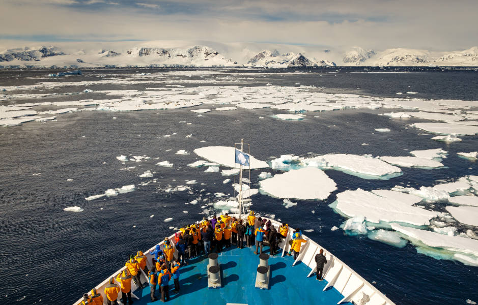 Guests in yellow parkas stand at the bow of their vessel as it cruises through sea ice in the Antarctic Peninsula.