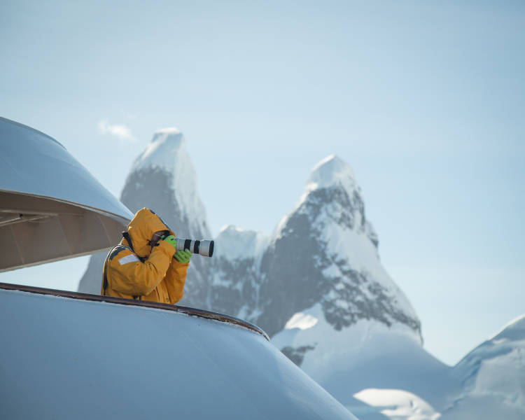 A guest in a yellow parka stands on one of the top decks of their vessel with a telephoto lens. Mountain peaks can be seen in the distance.