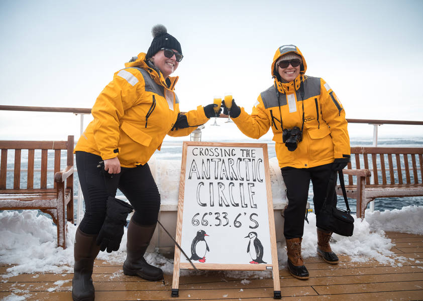 Two guests in yellow parkas pose for a photo next to a board that includes the coordinates of their ship, celebrating officially crossing the Antarctic Circle.