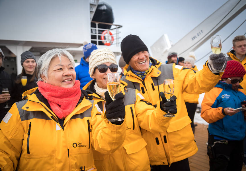 Three guests on yellow parkas raise their bubbly glasses as the Expedition Leader delivers their speech upon reaching the Antarctic Circle