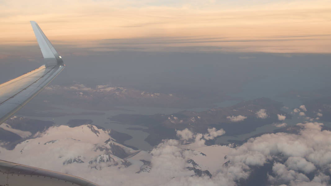 View from the window of an aircraft featuring its wing and a small cloud cover over the partially ice-covered peaks near Punta Arenas, Chile.