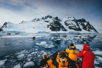 Guests in yellow parkas carefully watch an orca spyhopping as it tries to get better look at a seal resting on an ice flow.