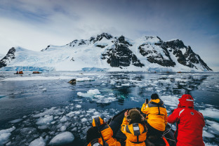 Guests in yellow parkas carefully watch an orca spyhopping as it tries to get better look at a seal resting on an ice flow.