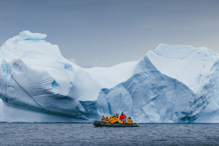 Zodiac boat in front of iceberg. Guests in yellow parkas can be seen taking photos while their Expedition Guide, in red, drives the boat.