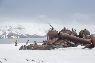 A guest in a yellow parka looks at the remnants of old and rusted equipment used to render fat from whale and seal blubber.