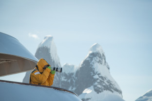 A guest in a yellow parka stands on one of the top decks of their vessel with a telephoto lens. Mountain peaks can be seen in the distance.