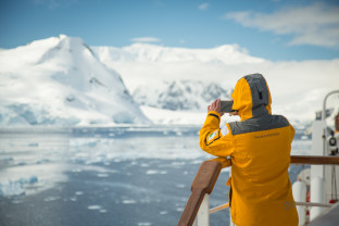 A guest in a yellow parka stands on one of the top decks of their vessel with a smartphone camera in hand.
