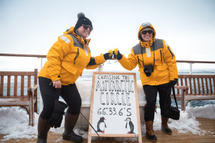 Two guests in yellow parkas raise their glasses to celebrate the achievement of Crossing the Antarctic Circle next to a sign that has the coordinates listed out.